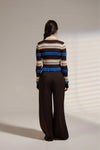 A model viewed from behind wearing a striped knit sweater with brown, beige, cream, and blue horizontal stripes, paired with dark brown wide-leg trousers, against a neutral studio backdrop with soft window shadow patterns.