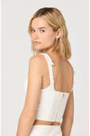 Woman shown from the back looking over her shoulder wearing a white sleeveless top with a ruffled back and back zipper on a plain background