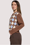 A woman modeling a brown long-sleeve sweater with an argyle pattern on the chest in brown, cream, and light blue, paired with brown trousers, against a solid light gray background.