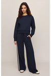 A woman modeling a matching navy blue lounge set consisting of a long-sleeve crewneck sweatshirt and wide-leg drawstring pants with white side stripes, set against a plain light beige studio background.