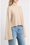 A woman wearing a light beige cropped knit sweater with dramatic bell sleeves and light blue jeans, photographed against a plain light gray studio background.