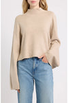 A woman wearing a beige knit mock-neck sweater with wide bell sleeves and light blue jeans, shown from the neck down against a plain light background.