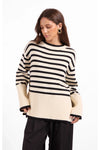 A woman wearing a cream and black horizontally striped knit sweater with ribbed cuffs and hem, paired with black pants, against a plain white studio background.