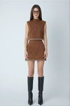 A model wearing a sleeveless brown faux-suede mini dress with a mock neck and a thin black and white patterned waistband, paired with black knee-high boots against a light gray studio background.