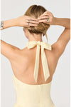 Woman shown from behind wearing a light yellow dress with a bow halter neck detail on a plain white background. 
