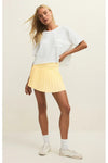 Woman wearing a white short sleeve loose top and a yellow pleated mini skirt with white crew socks and white sneakers on a plain background. 