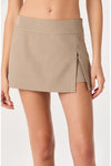 A model wearing a beige athletic mini skirt with a side slit, shown from the waist to the upper thighs against a light gray studio background.